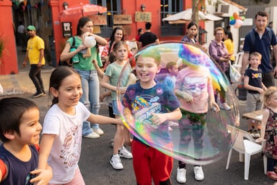 Niños sonrientes con camisetas coloridas, atrapando burbujas en un entorno festivo con adultos observando en un café colorido.