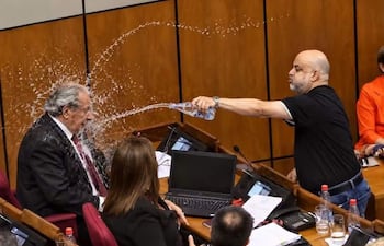 Hombre con camiseta negra arrojando agua mientras observan una mujer con abrigo negro y otros asistentes en un recinto cerrado.