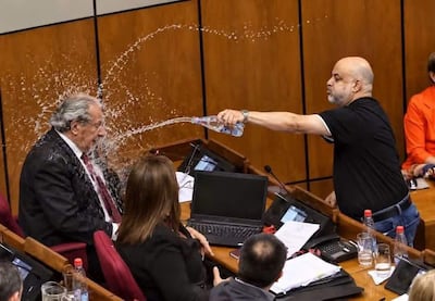 Hombre con camiseta negra arrojando agua mientras observan una mujer con abrigo negro y otros asistentes en un recinto cerrado.