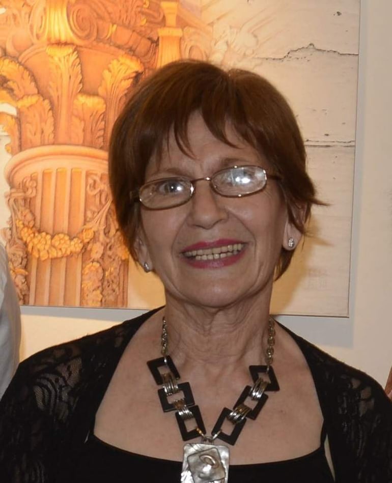 Mujer mayor sonriente con cabello castaño oscuro y gafas, lleva blusa negra y collar, en un ambiente elegante.