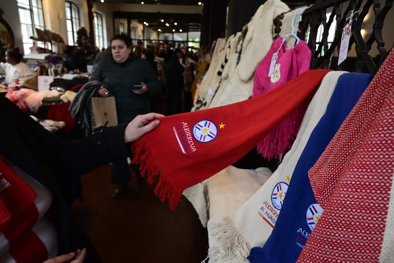 Mujer con chaqueta negra sostiene bufanda roja de la Albirroja en ambiente de feria, mientras otra mujer revisa su teléfono móvil.
