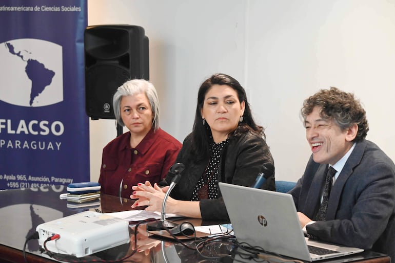 Panel con tres mujeres atentas y un hombre sonriente, micrófono y laptop sobre la mesa, logo de FLACSO al fondo.