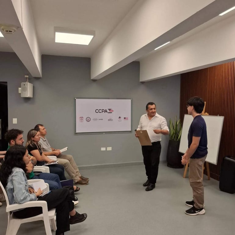 Hombre de pie con camisa blanca y carpeta, hablando a grupo que toma notas en sillas blancas en sala de reuniones moderna.
