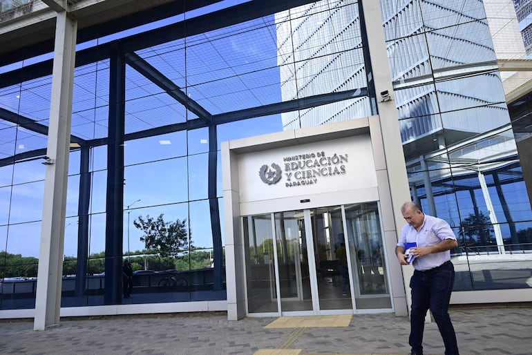 Hombre de pie junto a la entrada del Ministerio de Educación y Ciencias, sosteniendo un objeto y mirando hacia abajo en un entorno urbano.