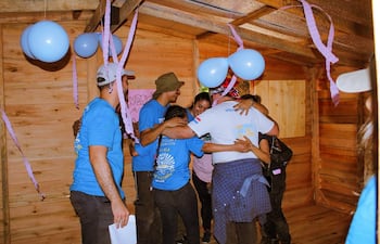 Siete personas con camisetas azules se abrazan en un ambiente festivo decorado con globos azules y cintas de papel.