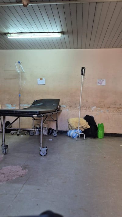 Sala de urgencias vacía con camilla negra, soporte de fluidos y bolsas en el suelo, paredes manchadas.