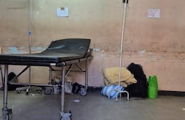 Sala de urgencias vacía con camilla negra, soporte de fluidos y bolsas en el suelo, paredes manchadas.