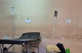 Sala de urgencias vacía con camilla negra, soporte de fluidos y bolsas en el suelo, paredes manchadas.
