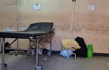 Sala de urgencias vacía con camilla negra, soporte de fluidos y bolsas en el suelo, paredes manchadas.