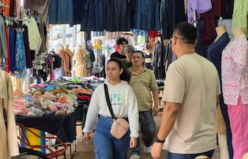 Multitud de compradores en un mercado exterior, con una mujer joven en abrigo blanco y hombres observando alrededor.