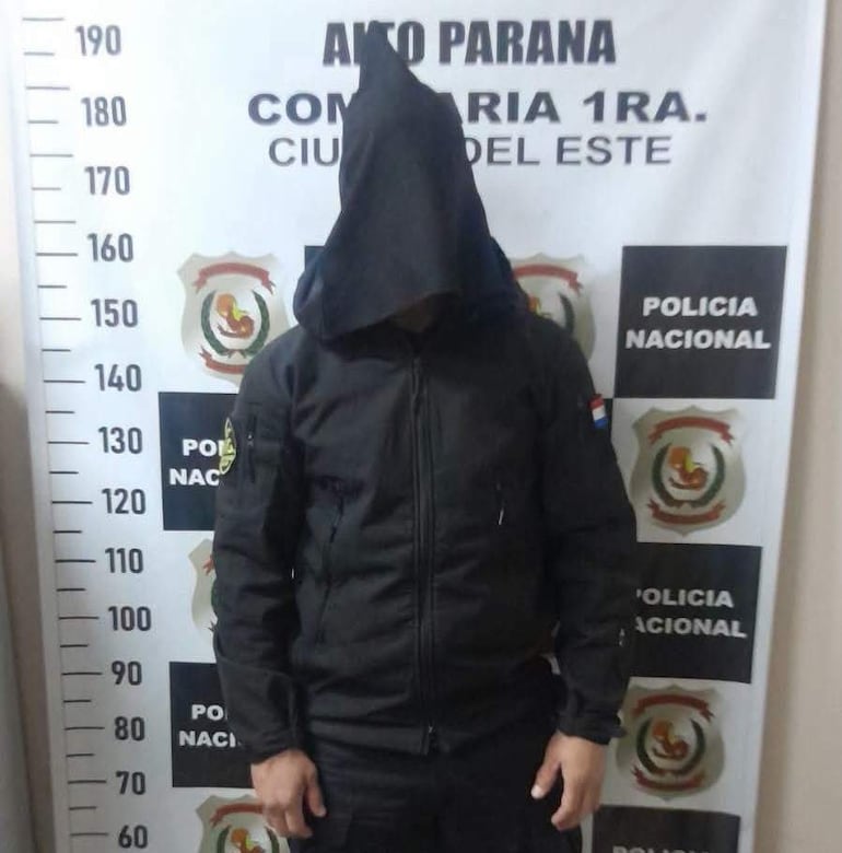 Persona con abrigo negro y capucha de pie frente a la Policía Nacional, en ambiente interior de comisaría.
