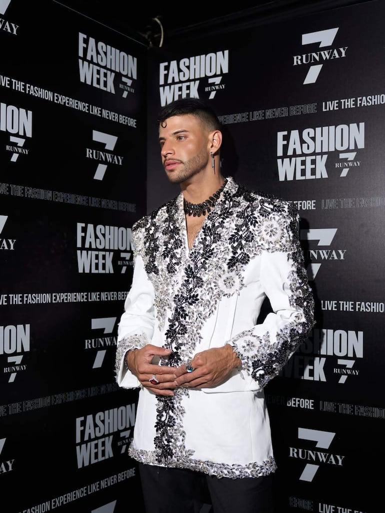 Hombre con chaqueta blanca brillante y detalles negros, posando en un evento de moda rodeado de carteles de Fashion Week.