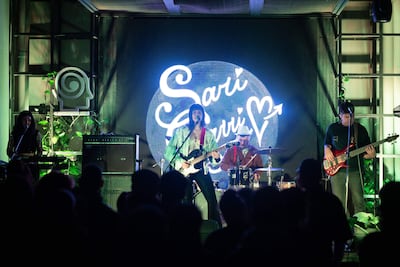 Mujer al frente con guitarra y micrófono, acompañada por músicos en un ambiente iluminado, durante el concierto de Sari Sari.