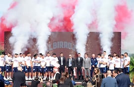 Futbolistas de Cerro Porteño en podio, vistiendo camisetas blancas con detalles institucionales, ambiente festivo con humo rojo y blanco.