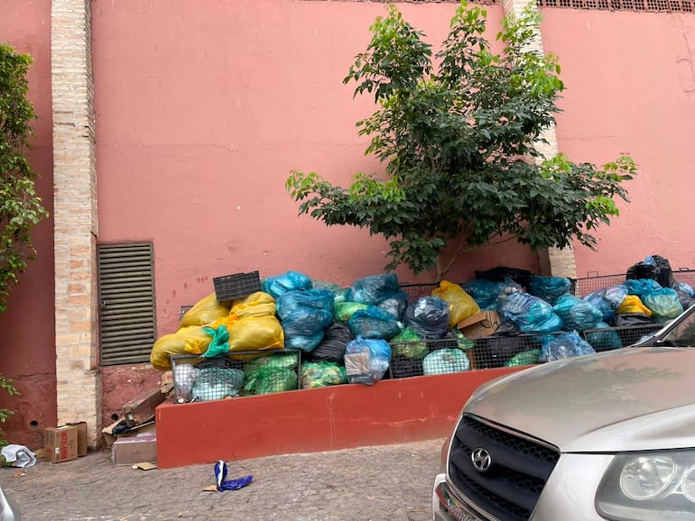 Acumulación de bolsas de basura de colores en una estructura de malla junto a una pared rosa, en un espacio público pavimentado.