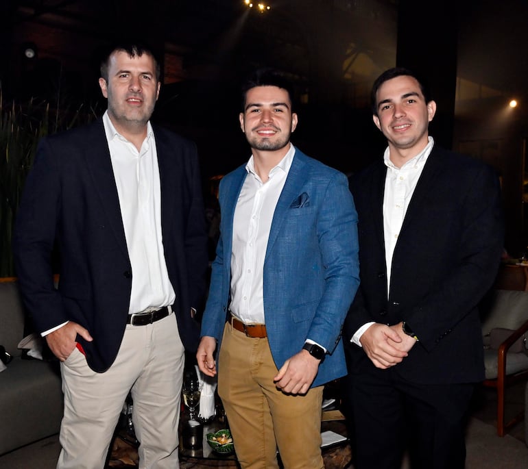 Tres hombres sonrientes: uno con saco oscuro, otro en chaqueta azul clara, y el tercero en saco negro, en un ambiente elegante.