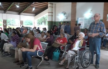 Mujer mayor en silla de ruedas junto a mujer de camiseta roja, varias personas esperando en sala informal.