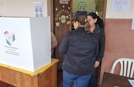 Mujer mayor con gafas es asistida por dos mujeres en un entorno de votación, una de ellas viste chaqueta negra y la otra abrigo claro.