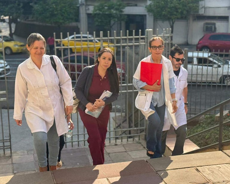 Mujeres en bata blanca y ropa de civiles suben escaleras, una de ellas lleva una carpeta roja, en un entorno urbano.