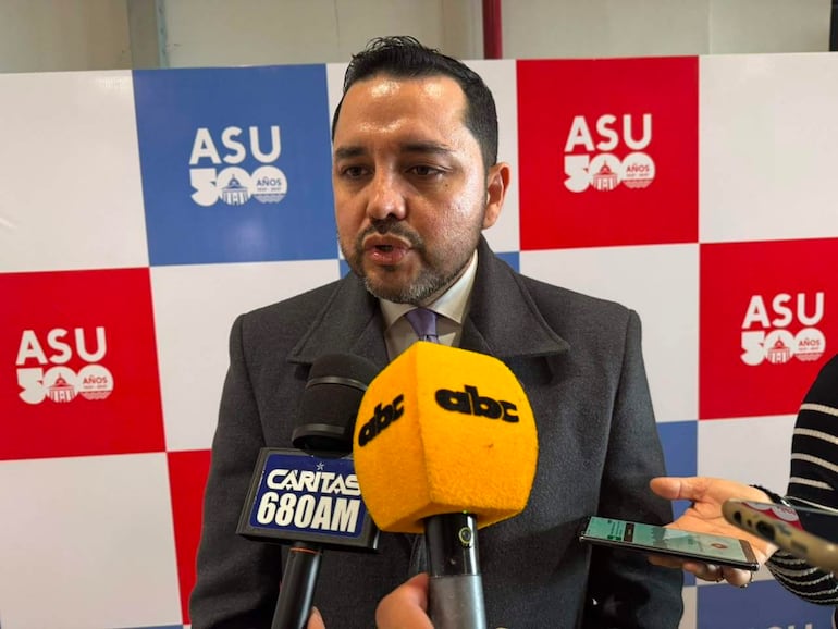 Luis Bello en chaqueta oscura y corbata clara habla al público, rodeado de micrófonos con logotipos de ABC Color y Cáritas 680 AM.