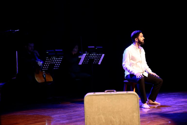 Diego Sánchez Haase sentado en un taburete con camisa blanca, rodeado de músicos con instrumentos en un teatro oscuro.