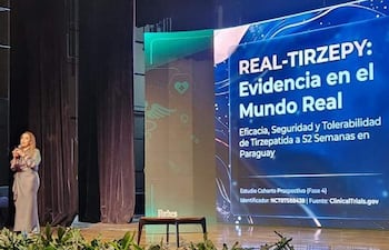 Dra. Elizabeth Valinotti Delmás de pie en un escenario, hablando con micrófono en un auditorio con decoración sencilla y pantalla detrás.