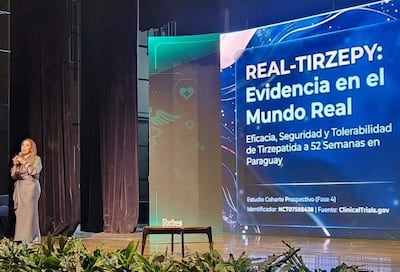 Dra. Elizabeth Valinotti Delmás de pie en un escenario, hablando con micrófono en un auditorio con decoración sencilla y pantalla detrás.