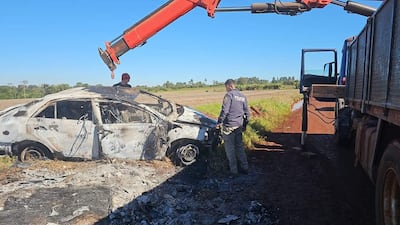 Oficial de policía examina vehículo incinerado, mientras un hombre en gorra roja opera un camión pesado en terreno rural.