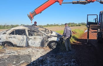 Oficial de policía examina vehículo incinerado, mientras un hombre en gorra roja opera un camión pesado en terreno rural.