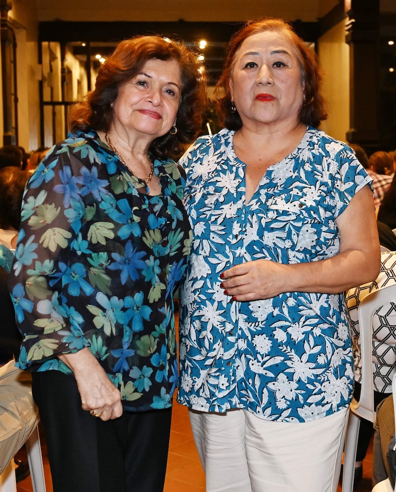 Mujeres de cabello castaño sonriendo. Una viste blusa oscura con flores azules y verdes, la otra blusa azul con flores blancas.