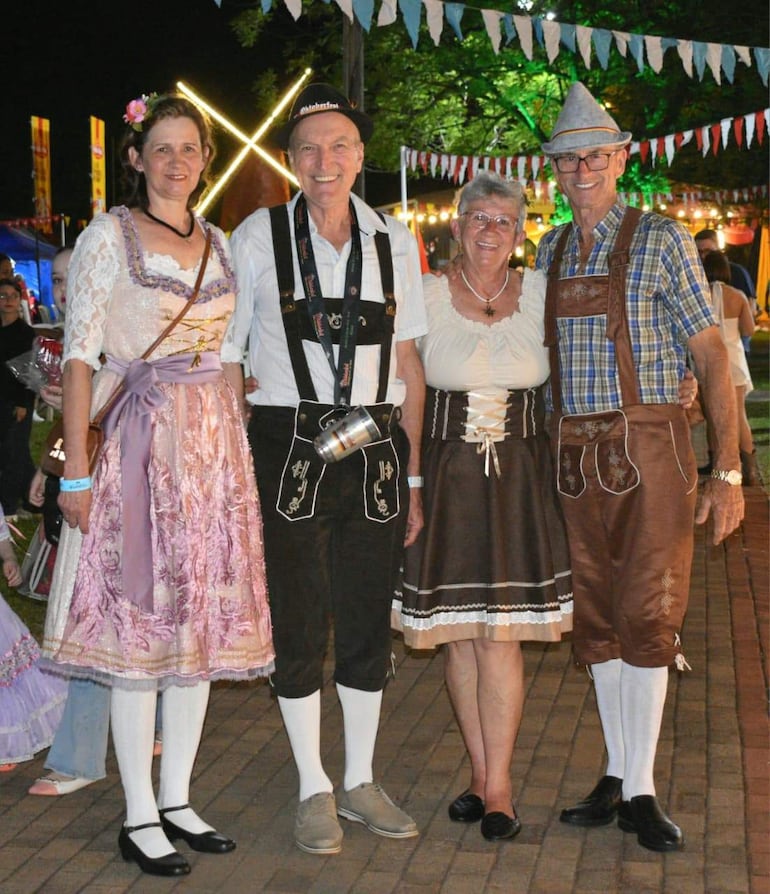 Cuatro personas sonrientes en trajes alemanes; mujer con vestido rosa, hombre con tirantes y jarra, mujer en blusa clara y falda oscura, hombre en camiseta de cuadros.