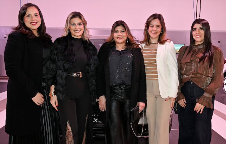Cinco mujeres sonrientes en grupo, con atuendos elegantes, destacando un abrigo negro en el centro.