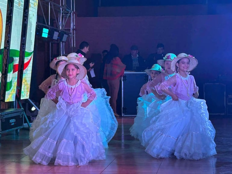 Niñas vestidas con trajes tradicionales, faldas blancas y blusas rosas, bailando con alegría en un ambiente iluminado.