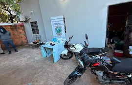 Dos motocicletas en primer plano; figura con chaqueta oscura observa mientras otra persona sujeta documentos frente a una puerta abierta.