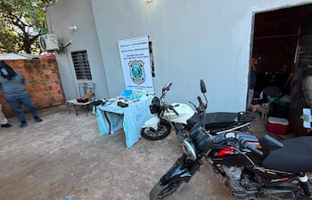 Dos motocicletas en primer plano; figura con chaqueta oscura observa mientras otra persona sujeta documentos frente a una puerta abierta.