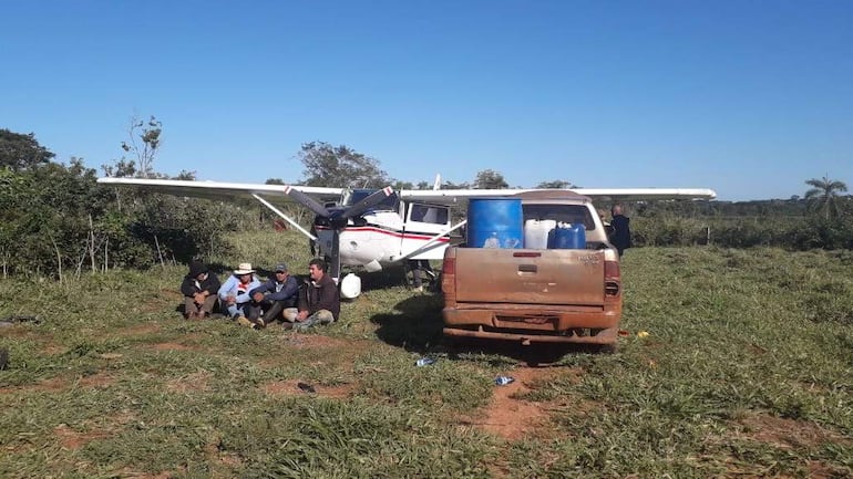 Seis individuos sentados en el suelo cerca de un avión blanco, con ropa de trabajo, un sombrero, y un vehículo pickup de fondo.