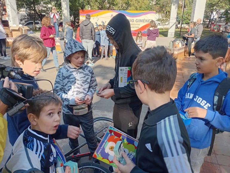 Grupo de niños interactuando en la plaza, algunos sosteniendo tarjetas, ambiente festivo y colorido.