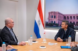 Emiliano Rolón Fernández en traje oscuro, junto a Santiago Peña Palacios en perfil, en oficina con bandera paraguaya de fondo.