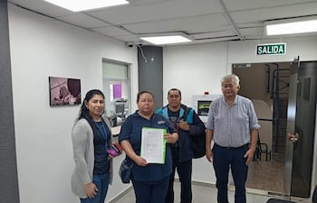Mujeres en camisetas oscuras sosteniendo documentos, hombre canoso observa, ambiente de oficina moderno con cartel de salida.