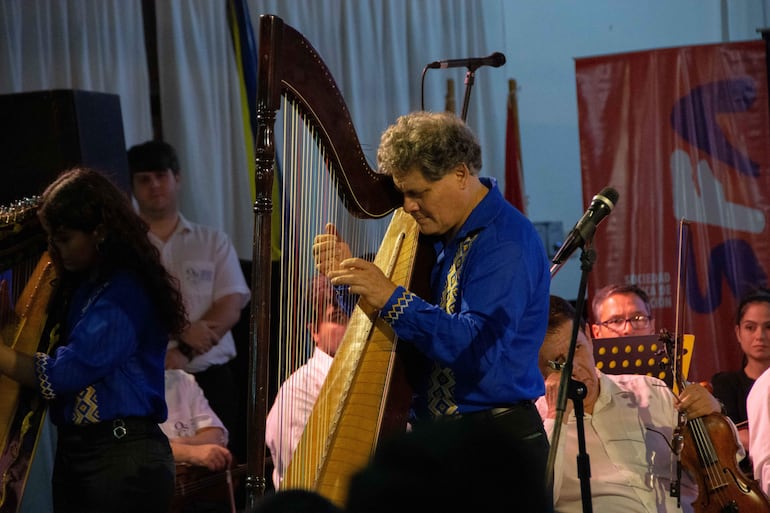 Marcos Lucena, con cabello rizado y gafas, toca el arpa y lleva camisa azul, mientras otros músicos lo acompañan en el auditorio.