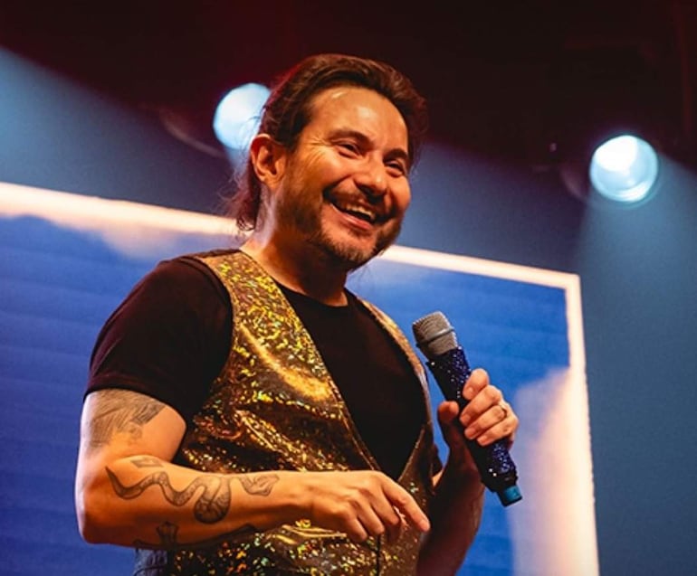 Felipe Avello en el escenario, con camiseta negra y chaleco brillante, sonríe mientras sostiene un micrófono en una atmósfera festiva.