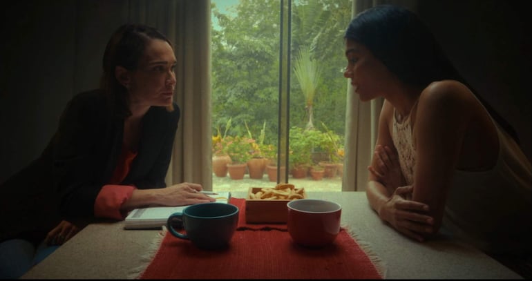 Lali González con saco negro y blusa roja, hablando con otra mujer de blusa blanca, en mesa con tazas y galletas.