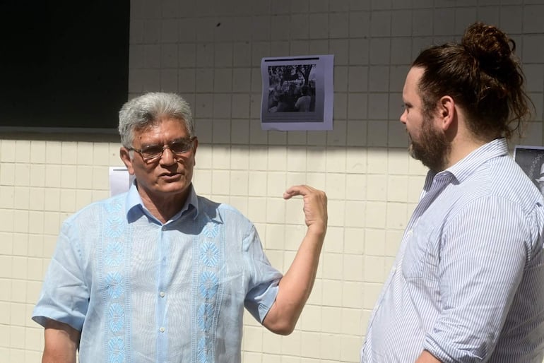 Hombre mayor con gafas y camisa clara gesticula mientras habla con un hombre más joven de cabello rizado y camisa a rayas.