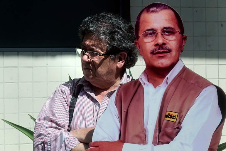 Hombre de cabello oscuro y rizado en camisa de rayas escucha atentamente, mientras otro hombre con chaleco beige con logo de ABC Color está presente.