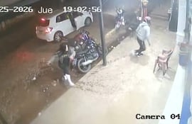 Una mujer con abrigo gris y botas blancas se mueve, gesticulando, mientras una persona camina a su lado en un ambiente urbano nocturno.