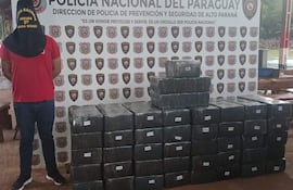 Hombre de espaldas con camiseta roja y pantalón de mezclilla, junto a paquetes de marihuana en un entorno policial.