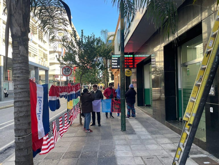 Grupo de cinco personas en acera, conversando y observando. Banderas y camisetas colgadas, ambiente festivo.