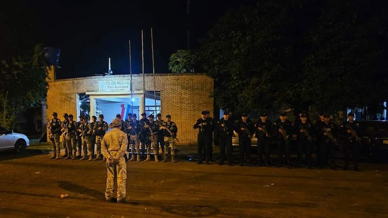 Quince agentes de policía en uniformes oscuros alineados, algunos armados, frente a la comisaría María Auxiliadora.