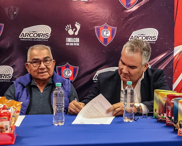 Hombre mayor con chaleco azul sonríe, mientras joven con traje oscuro sostiene documentos en la sala de prensa del Club Cerro Porteño.