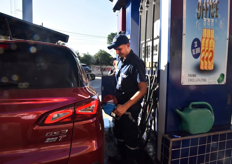 Empleado con camiseta oscura y sombrero sostiene manguera en estación de servicio, con fondo de graffitis y publicidad.